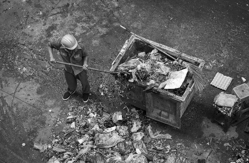A person wearing a protective helmet and work clothes is shown standing on a paved or gravel surface outdoors, using a long-handled tool to clear a pile of mixed construction and garden waste from the ground into a large, open-top waste skip. The skip, positioned centrally in the image, is filled with various debris including wood, plastic, and organic material. To the right of the skip, a small stack of bricks or paving slabs is visible, alongside some loose materials and a nearby container or toolbox. The scene appears to be part of a garden clearance operation, possibly in a residential area, with natural daylight illuminating the area. The surroundings suggest a driveway or outdoor workspace typical for rubbish removal services like those offered by Garden Clearance Pinner, servicing areas within postcode regions such as Pinner or nearby London suburbs. The focus is on the manual loading of waste into the skip, with a clear environment dedicated to professional rubbish disposal.