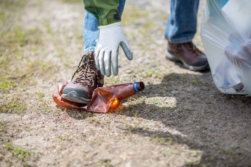 A person wearing blue jeans, brown working boots, and white gloves is picking up a discarded glass bottle from a dirt pathway, with a partially filled grey rubbish bag nearby. The bottle has an amber colour and is lying on the uneven ground, which has patches of grass and soil. In the background, a plastic rubbish bag and a leaflet or paper are visible, indicating a cleanup effort. This scene occurs outdoors, possibly in a garden or park area within or near Pinner, where Garden Clearance Pinner provides rubbish removal services. The image emphasizes cleanliness and waste collection in a natural setting, highlighting the importance of proper rubbish disposal.