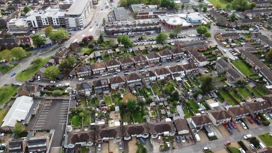 An aerial view of a residential neighbourhood with a row of semi-detached houses, each featuring small, well-maintained gardens with grass lawns, trees, and garden furniture. The houses have brick ext