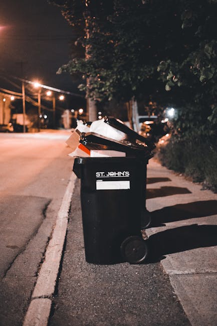 Side view of a small, old blue pickup truck parked on a city street, loaded heavily with various types of rubbish including cardboard boxes, wooden pallets, black plastic bags, and loose debris. The t